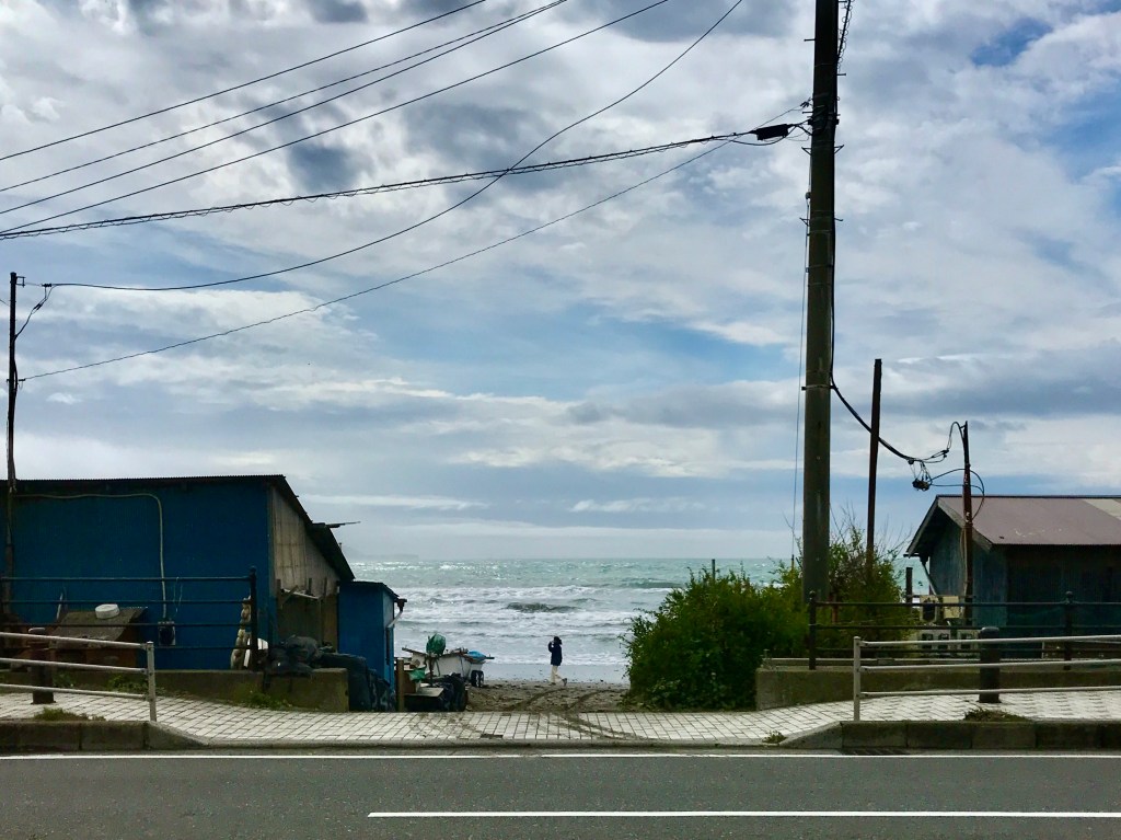 The bay seen between two fishing huts, with a single person walking along the beach.